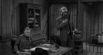 Movie still from “Terror in a Texas Town” (1958), directed by Joseph H. Lewis – Two men are sitting at a desk and one is holding a hat; Medium shot, Over the shoulder angle