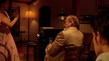 Movie still from “Tetro” (2009), directed by Francis Ford Coppola – A man sitting in front of a piano holding a glass of wine; Medium shot, Over the shoulder angle