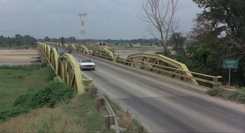 Movie still from “Tex” (1982), directed by Tim Hunter – A car is driving down a road with a bridge in the background; Extreme Wide shot, High angle