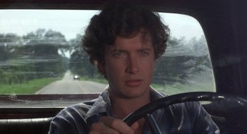 Movie still from “Tex” (1982), directed by Tim Hunter – A man sitting in the drivers seat of a car driving down a road; Close Up shot, Over the shoulder angle