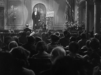 Movie still from “The 39 Steps” (1935), directed by Alfred Hitchcock – An old photo of a crowd of people watching a man on a stage; Extreme Wide shot, Low angle