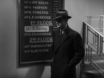 Movie still from “The 39 Steps” (1935), directed by Alfred Hitchcock – A black and white photo of a man standing in front of a sign; Medium shot, High angle
