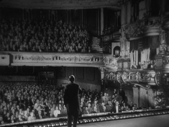 Movie still from “The 39 Steps” (1935), directed by Alfred Hitchcock – A man standing on a stage in front of an audience; Extreme Wide shot, High angle