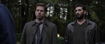 Movie still from “Department Q: The Absent One” (2014), directed by Mikkel Nørgaard – Two men standing next to each other in front of trees; Medium shot, Over the shoulder angle