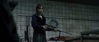 Movie still from “Department Q: The Absent One” (2014), directed by Mikkel Nørgaard – A person holding a rifle in a room; Medium shot, Over the shoulder angle