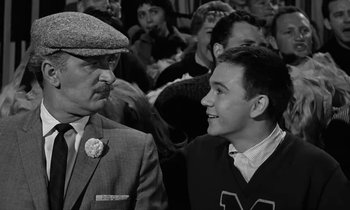 Movie still from “The Absent Minded Professor” (1961), directed by Robert Stevenson – A man in a suit and a man in a flat cap talking to another man in front of a crowd of onlookers; Close Up shot, Over the shoulder angle