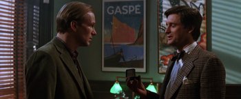 Movie still from “The Accidental Tourist” (1988), directed by Lawrence Kasdan – Two men are talking in front of a poster of gaspé; Medium shot, Over the shoulder angle