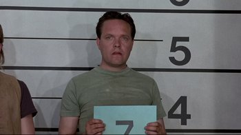 Movie still from “The Accused” (1988), directed by Jonathan Kaplan – A man holding up a sign in front of a mugshot wall; Close Up shot, Low angle