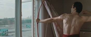 Movie still from “The Adjuster” (1991), directed by Atom Egoyan – A man holding a bow and arrow in a room; Medium shot, Low angle