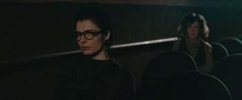 Movie still from “The Adjuster” (1991), directed by Atom Egoyan – A woman wearing glasses sitting in front of a wall; Close Up shot, Low angle