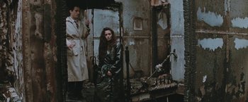 Movie still from “The Adjuster” (1991), directed by Atom Egoyan – A man and a woman standing next to each other in front of a fire damaged building; Wide shot, High angle