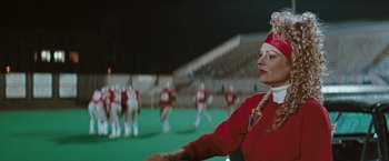 Movie still from “The Adjuster” (1991), directed by Atom Egoyan – A woman in a red uniform stands in front of a group of people in a stadium; Medium shot, Low angle