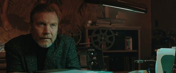 Movie still from “The Adjuster” (1991), directed by Atom Egoyan – A man sitting at a table in front of an old movie projector; Close Up shot, Low angle