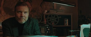 Movie still from “The Adjuster” (1991), directed by Atom Egoyan – A man sitting at a table in front of a projector; Close Up shot, Low angle