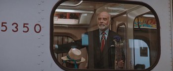 Movie still from “The Adjuster” (1991), directed by Atom Egoyan – An older man in a suit and tie on a train; Medium shot, Over the shoulder angle