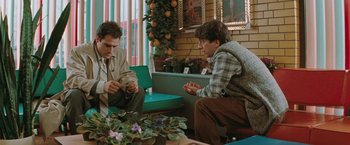 Movie still from “The Adjuster” (1991), directed by Atom Egoyan – Two men sitting at a table in front of potted plants; Medium shot, Over the shoulder angle