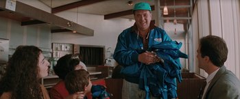 Movie still from “The Adjuster” (1991), directed by Atom Egoyan – An older man wearing a baseball cap and a blue rain jacket; Medium shot, Over the shoulder angle