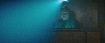 Movie still from “The Adjuster” (1991), directed by Atom Egoyan – A man sitting at a desk in a dark room; Medium shot, Low angle