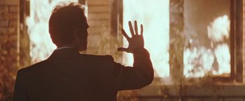 Movie still from “The Adjuster” (1991), directed by Atom Egoyan – A man in a black suit is holding his hand up to a window; Medium shot, Over the shoulder angle