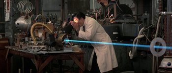 Movie still from “The Adventures of Buckaroo Banzai Across the 8th Dimension” (1984), directed by W.D. Richter – A man in a white lab coat looking through a microscope; Wide shot, High angle
