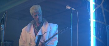 Movie still from “The Adventures of Buckaroo Banzai Across the 8th Dimension” (1984), directed by W.D. Richter – A man in a white jacket is playing a guitar; Medium shot, Low angle