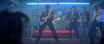 Movie still from “The Adventures of Buckaroo Banzai Across the 8th Dimension” (1984), directed by W.D. Richter – A man in a suit and tie playing an electric guitar in front of a microphone; Wide shot, Low angle