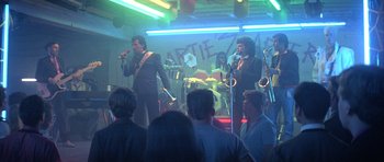 Movie still from “The Adventures of Buckaroo Banzai Across the 8th Dimension” (1984), directed by W.D. Richter – Two men in suits singing and playing instruments in front of an audience; Wide shot, Over the shoulder angle
