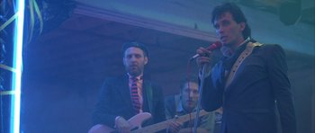Movie still from “The Adventures of Buckaroo Banzai Across the 8th Dimension” (1984), directed by W.D. Richter – A man in a suit and tie playing a guitar and a microphone; Medium shot, Low angle