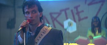 Movie still from “The Adventures of Buckaroo Banzai Across the 8th Dimension” (1984), directed by W.D. Richter – A man in a suit standing in front of a microphone; Close Up shot, Low angle