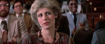 Movie still from “The Adventures of Buckaroo Banzai Across the 8th Dimension” (1984), directed by W.D. Richter – An older woman wearing a necklace in front of a group of people; Close Up shot, Over the shoulder angle