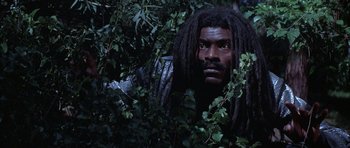Movie still from “The Adventures of Buckaroo Banzai Across the 8th Dimension” (1984), directed by W.D. Richter – A man with dreadlocks is hiding in the bushes; Medium shot, Low angle