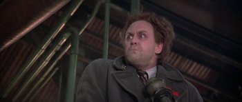 Movie still from “The Adventures of Buckaroo Banzai Across the 8th Dimension” (1984), directed by W.D. Richter – A man in a gray coat and a microphone; Close Up shot, Low angle