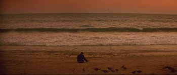 Movie still from “The Adventures of Ford Fairlane” (1990), directed by Renny Harlin – A man sitting on the beach watching the sun go down over the ocean; Extreme Wide shot, High angle