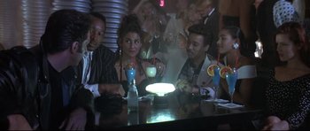 Movie still from “The Adventures of Ford Fairlane” (1990), directed by Renny Harlin – A group of people sitting around a table with drinks; Medium shot, Over the shoulder angle