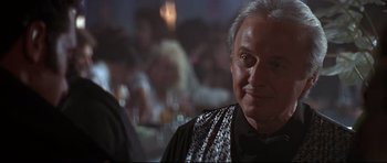 Movie still from “The Adventures of Ford Fairlane” (1990), directed by Renny Harlin – An older man wearing a black shirt and a black and white scarf; Close Up shot, Over the shoulder angle