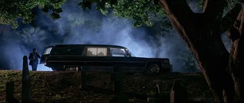Movie still from “The Adventures of Ford Fairlane” (1990), directed by Renny Harlin – A hearse is parked in a wooded area near trees; Wide shot, Low angle