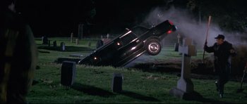 Movie still from “The Adventures of Ford Fairlane” (1990), directed by Renny Harlin – A car that is falling off of the ground in a cemetery; Wide shot, High angle