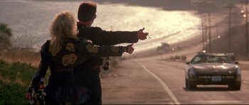 Movie still from “The Adventures of Ford Fairlane” (1990), directed by Renny Harlin – A man standing on the side of a road holding a cigarette; Wide shot, Low angle