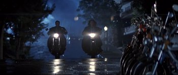 Movie still from “The Adventures of Ford Fairlane” (1990), directed by Renny Harlin – Two people riding motorcycles down a street at night; Wide shot, Low angle