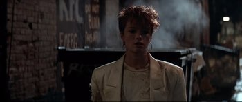 Movie still from “The Adventures of Ford Fairlane” (1990), directed by Renny Harlin – A woman in a white jacket standing next to a building; Medium shot, Low angle