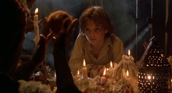 Movie still from “The Adventures of Huck Finn” (1993), directed by Stephen Sommers – A young boy sitting in front of a table of candles; Medium shot, Over the shoulder angle