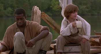 Movie still from “The Adventures of Huck Finn” (1993), directed by Stephen Sommers – Two people sitting on a dock near a body of water; Medium shot, Over the shoulder angle