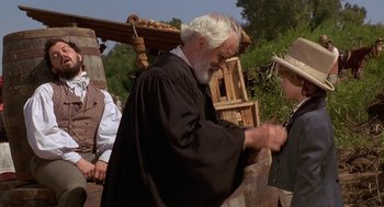 Movie still from “The Adventures of Huck Finn” (1993), directed by Stephen Sommers – An old man in a black robe is tying a hat; Medium shot, Over the shoulder angle