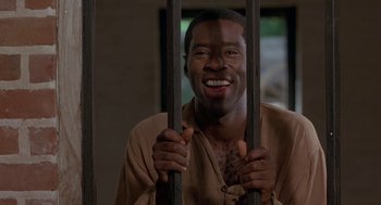 Movie still from “The Adventures of Huck Finn” (1993), directed by Stephen Sommers – A man holding onto bars and smiling for the camera; Close Up shot, High angle