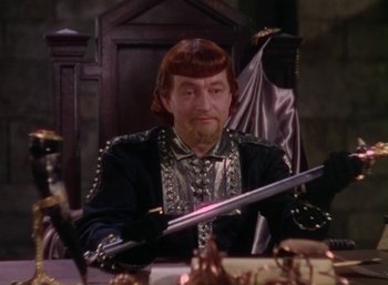 Movie still from “The Adventures of Robin Hood” (1938), directed by William Keighley – A man sitting at a table holding a sword in front of him; Medium shot, High angle