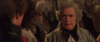 Movie still from “The Affair of the Necklace” (2001), directed by Charles Shyer – An older man in a wig and a wig stand in front of other people; Close Up shot, Over the shoulder angle