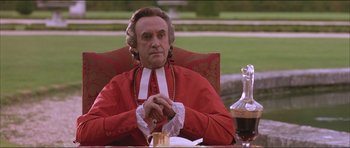 Movie still from “The Affair of the Necklace” (2001), directed by Charles Shyer – A man sitting in a chair in front of a bottle of wine; Medium shot, Over the shoulder angle