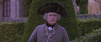 Movie still from “The Affair of the Necklace” (2001), directed by Charles Shyer – An old man in a wig and a hat is standing in front of a hedge; Close Up shot, Low angle