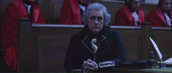 Movie still from “The Affair of the Necklace” (2001), directed by Charles Shyer – An older man sitting in a courtroom holding his hands together; Medium shot, High angle