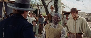 Movie still from “The Alamo” (1960), directed by John Wayne – An older man in a hat and a crowd of people; Medium shot, Over the shoulder angle